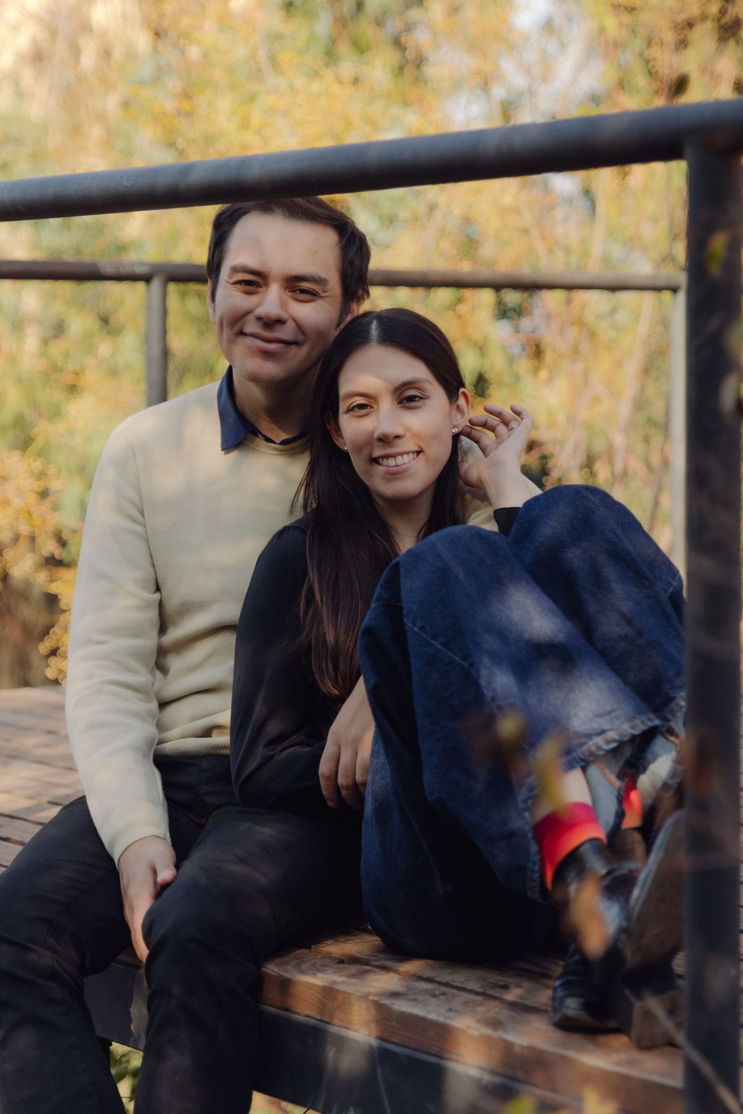 Smiling couple sitting together on a wooden bench in an autumn park, man in cream sweater embracing woman in black top.