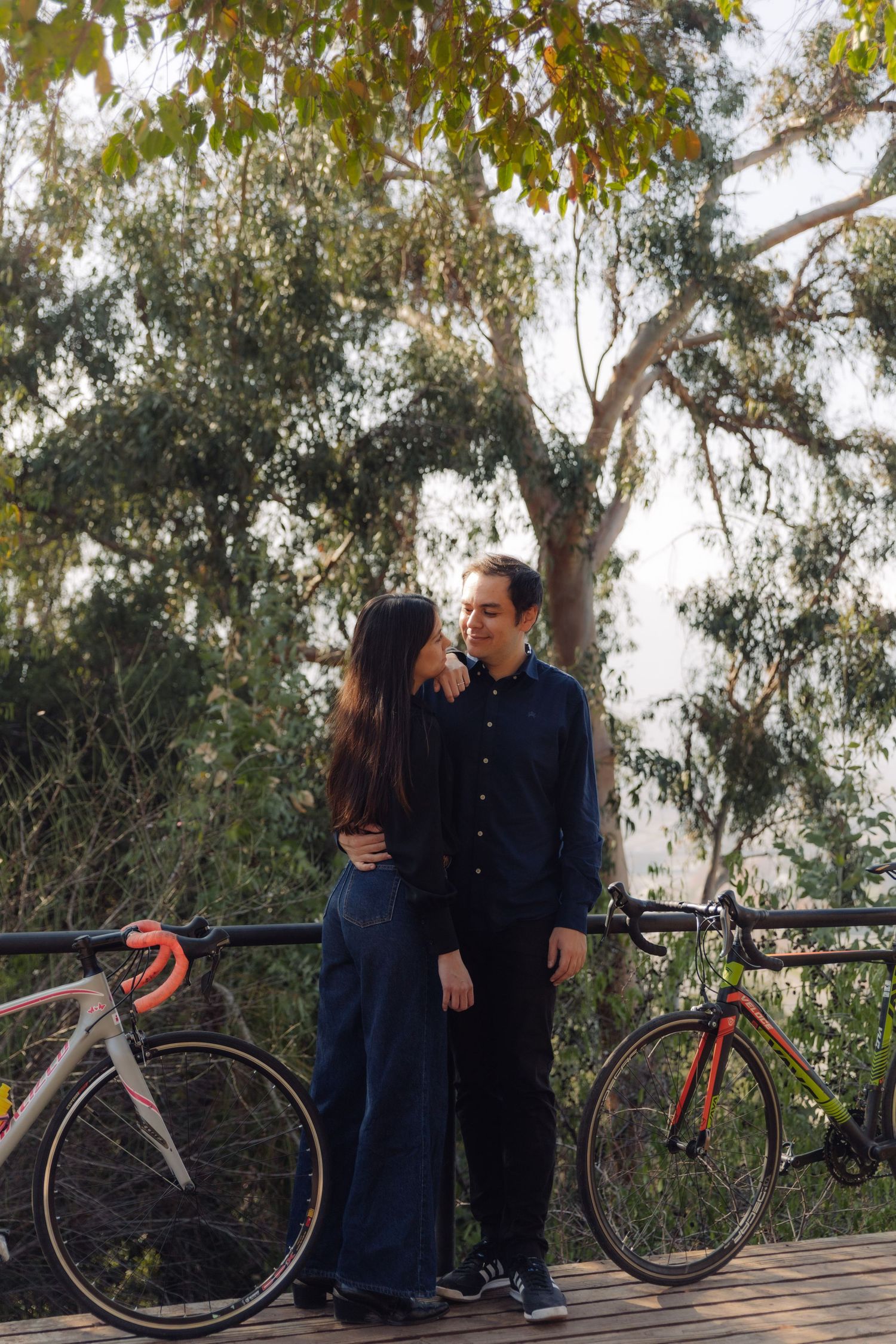 Couple standing close together on a wooden deck surrounded by trees, with two road bicycles leaning on a railing.