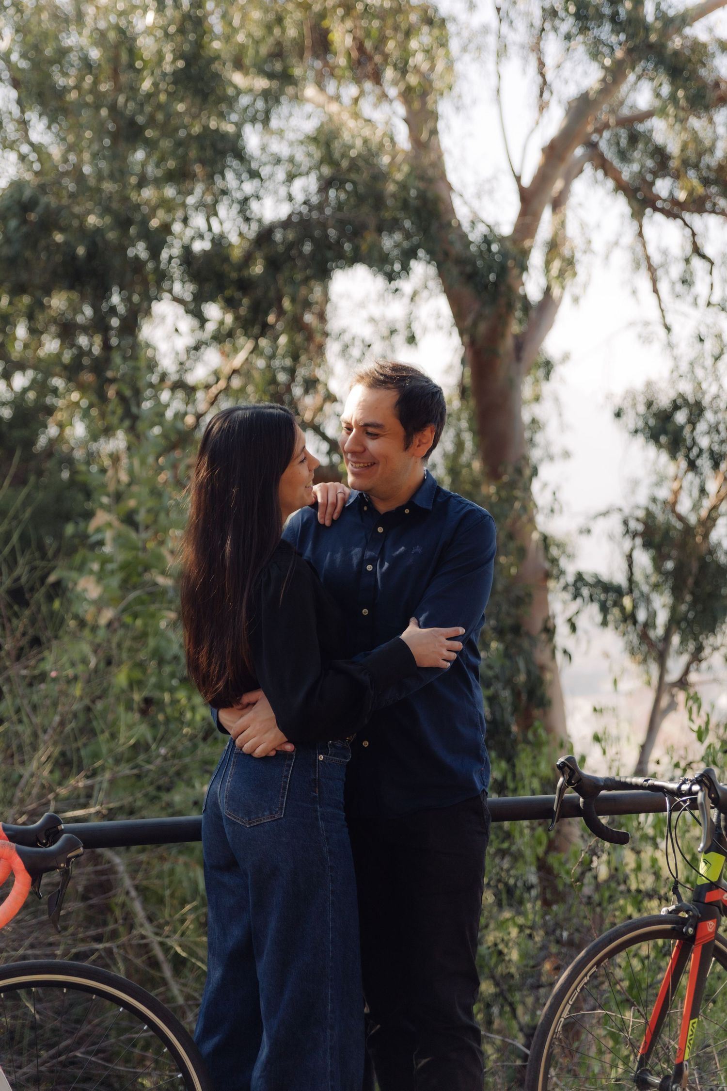 Happy couple embracing and smiling at each other outdoors, with colorful road bikes and lush green trees behind them.