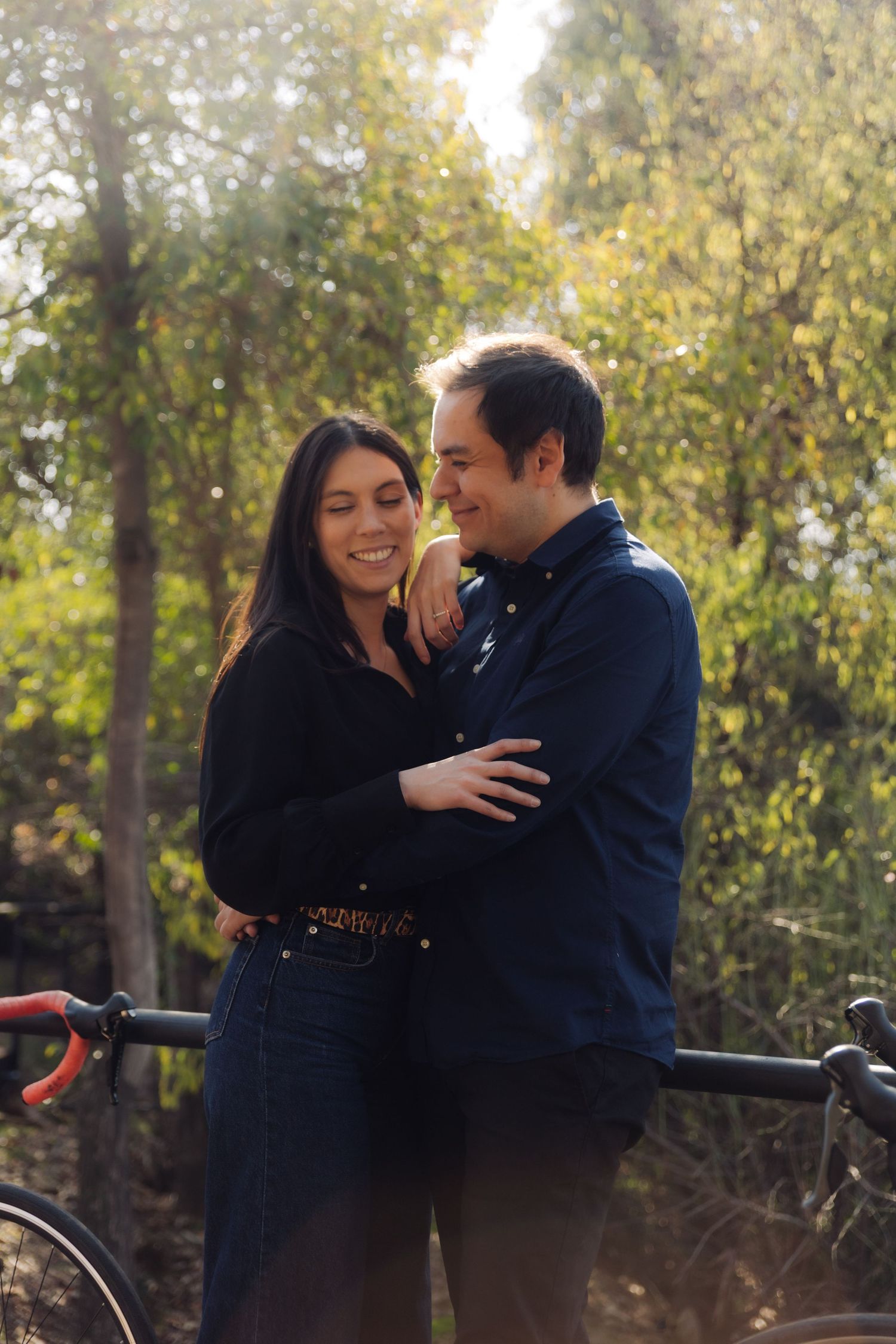 Laughing couple hugging near road bicycles in a sunlit park, woman in black shirt and jeans enjoying a joyful moment.