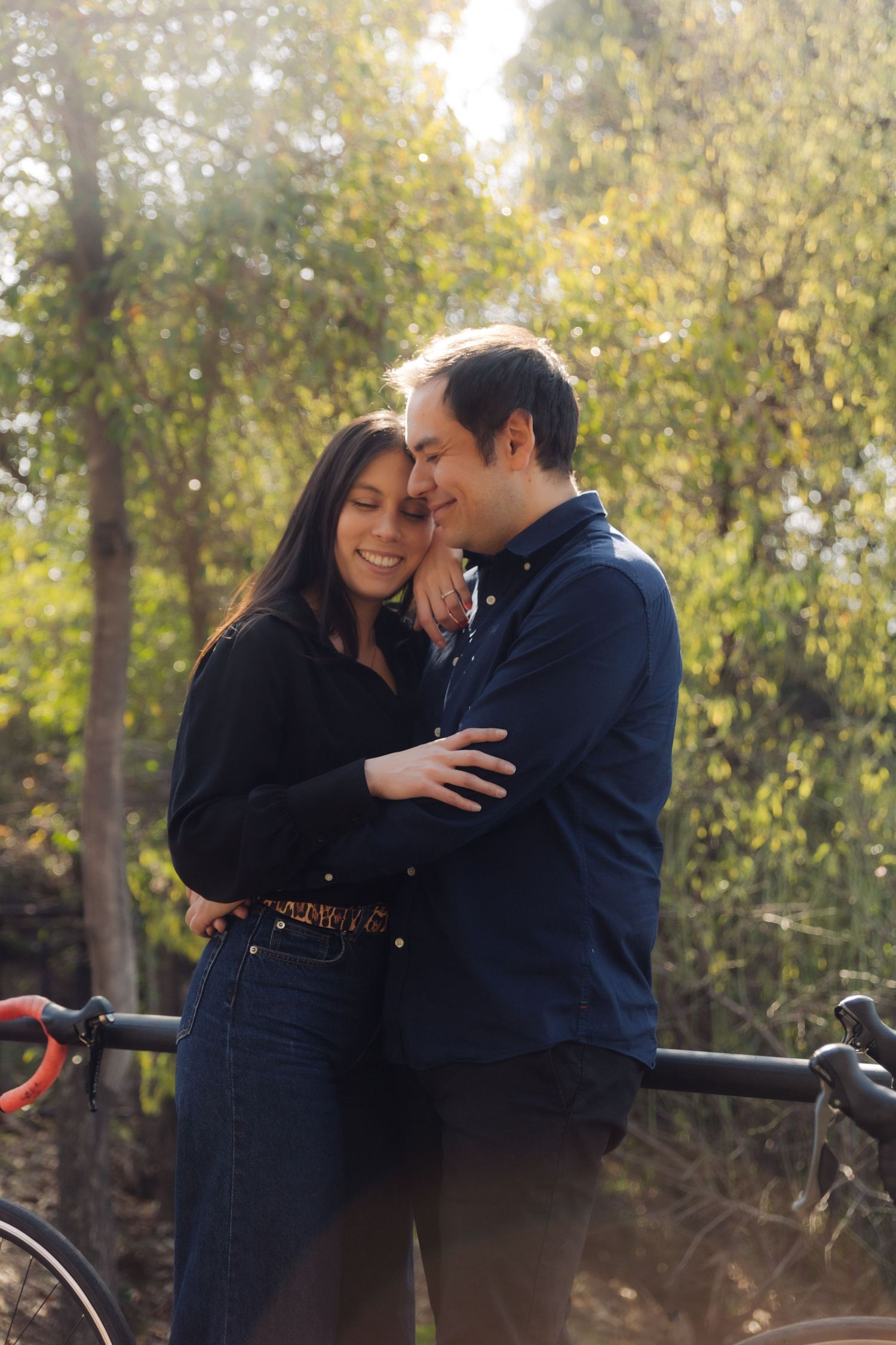 Couple sharing a romantic embrace in a sun-dappled park, standing close together with bicycles resting on a railing.