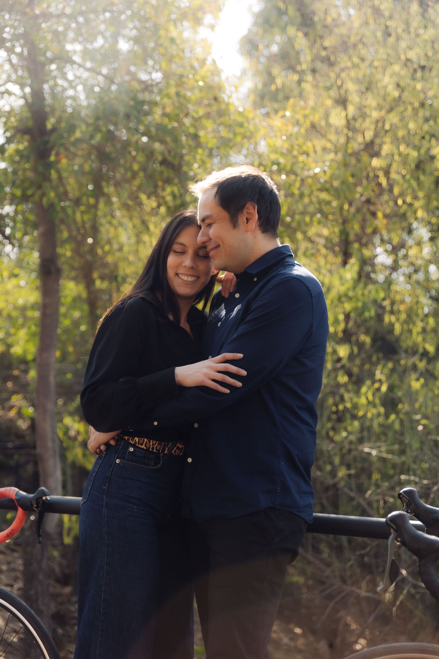 Couple cuddling closely in a bright outdoor park setting, smiling together with bicycles and green trees in background.