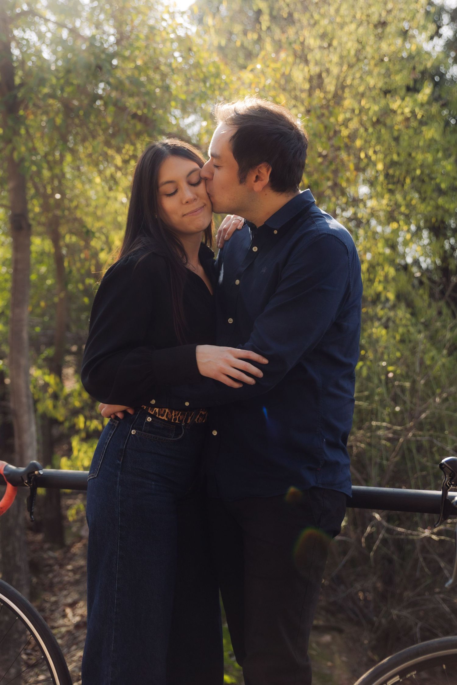 Man kissing woman on the cheek outdoors in golden sunlight, both standing near road bikes with lush trees behind them.