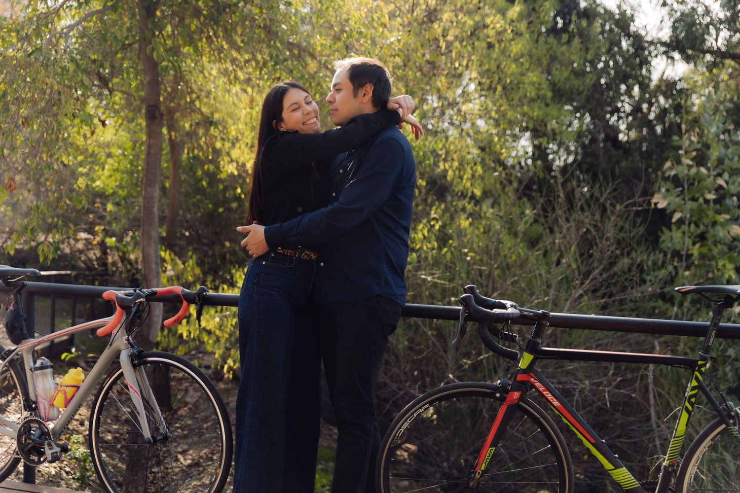 Couple sharing a kiss while embracing near two road bicycles on a tree-lined path in a peaceful outdoor setting.