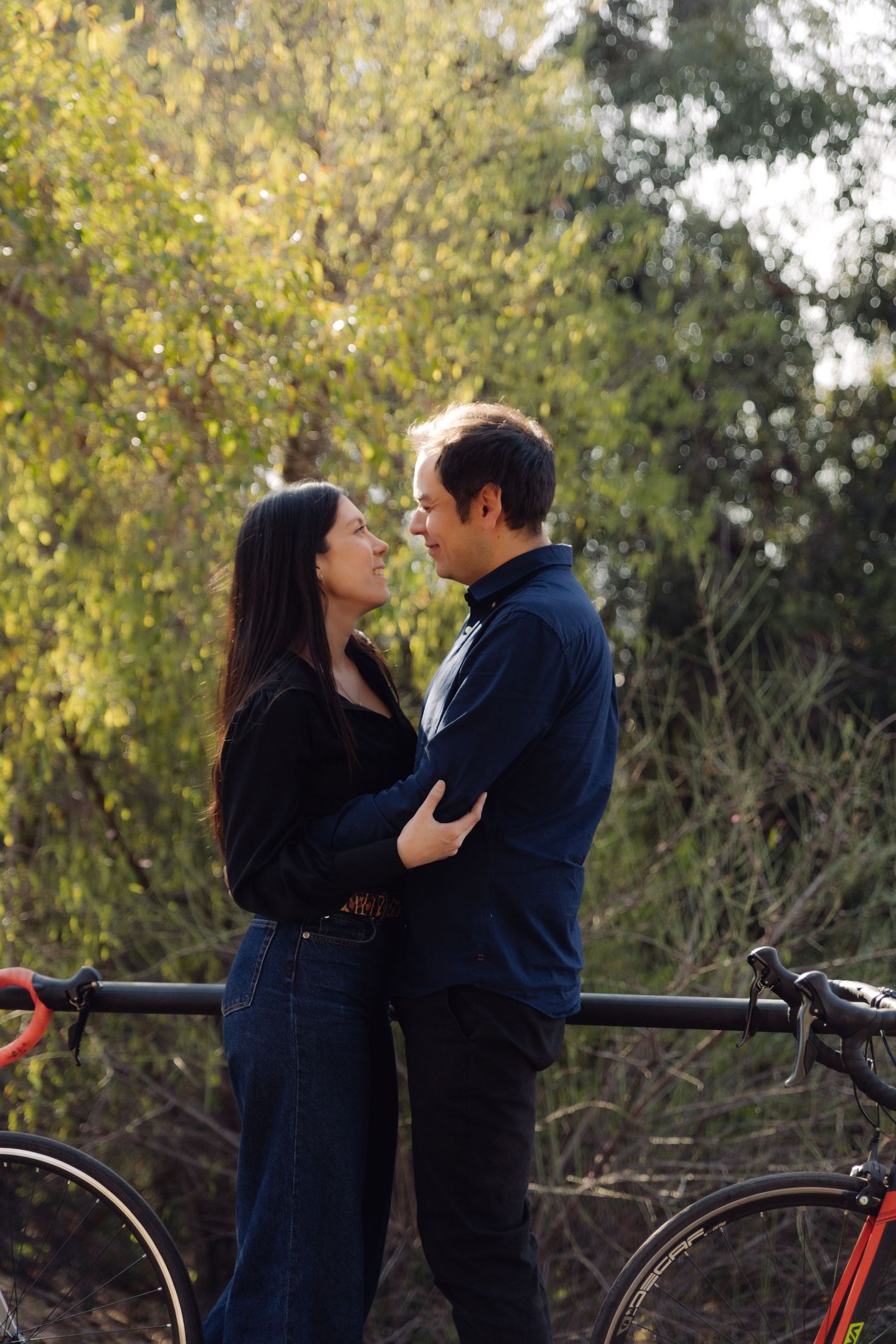 Couple gazing into each other eyes while embracing outdoors, standing beside road bikes with vibrant autumn foliage behind.
