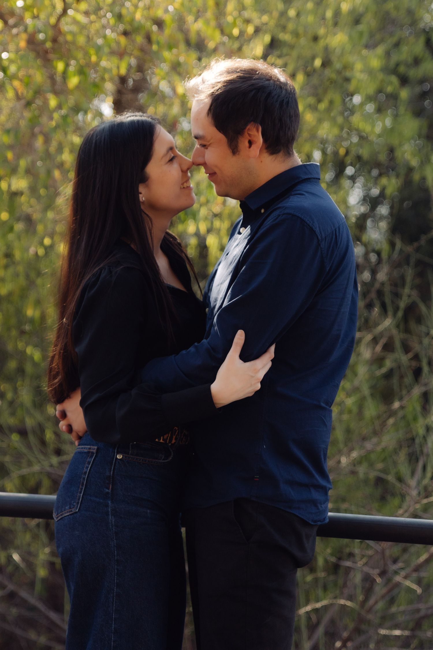 Romantic couple almost kissing while embracing outdoors near a railing with green trees in background.