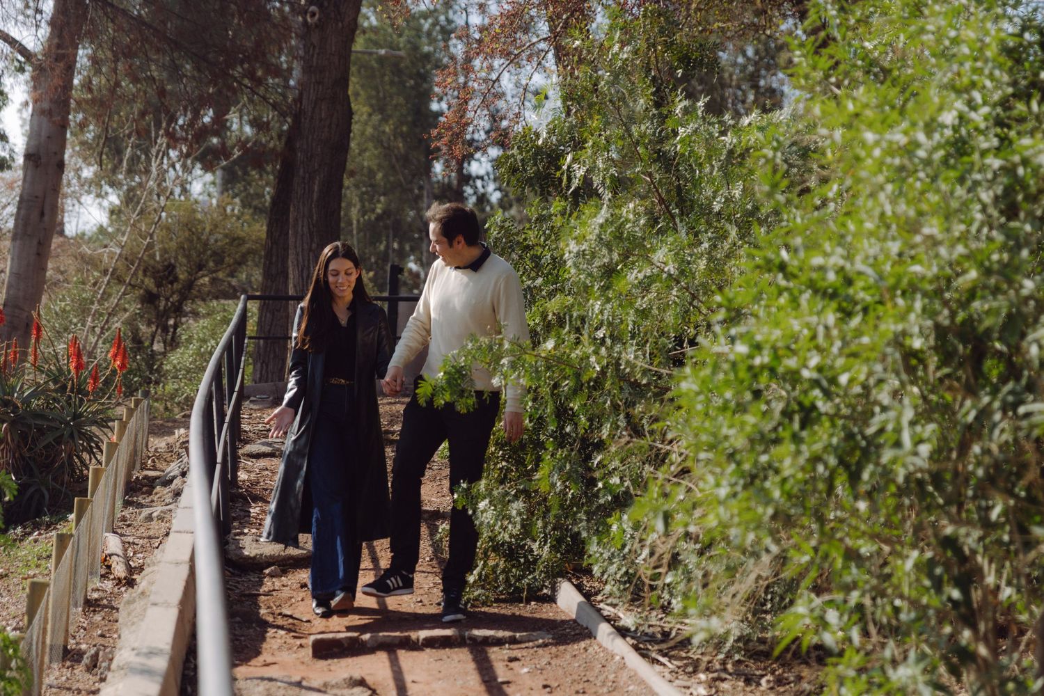 Couple walking hand in hand along a sunny garden path surrounded by lush green trees.