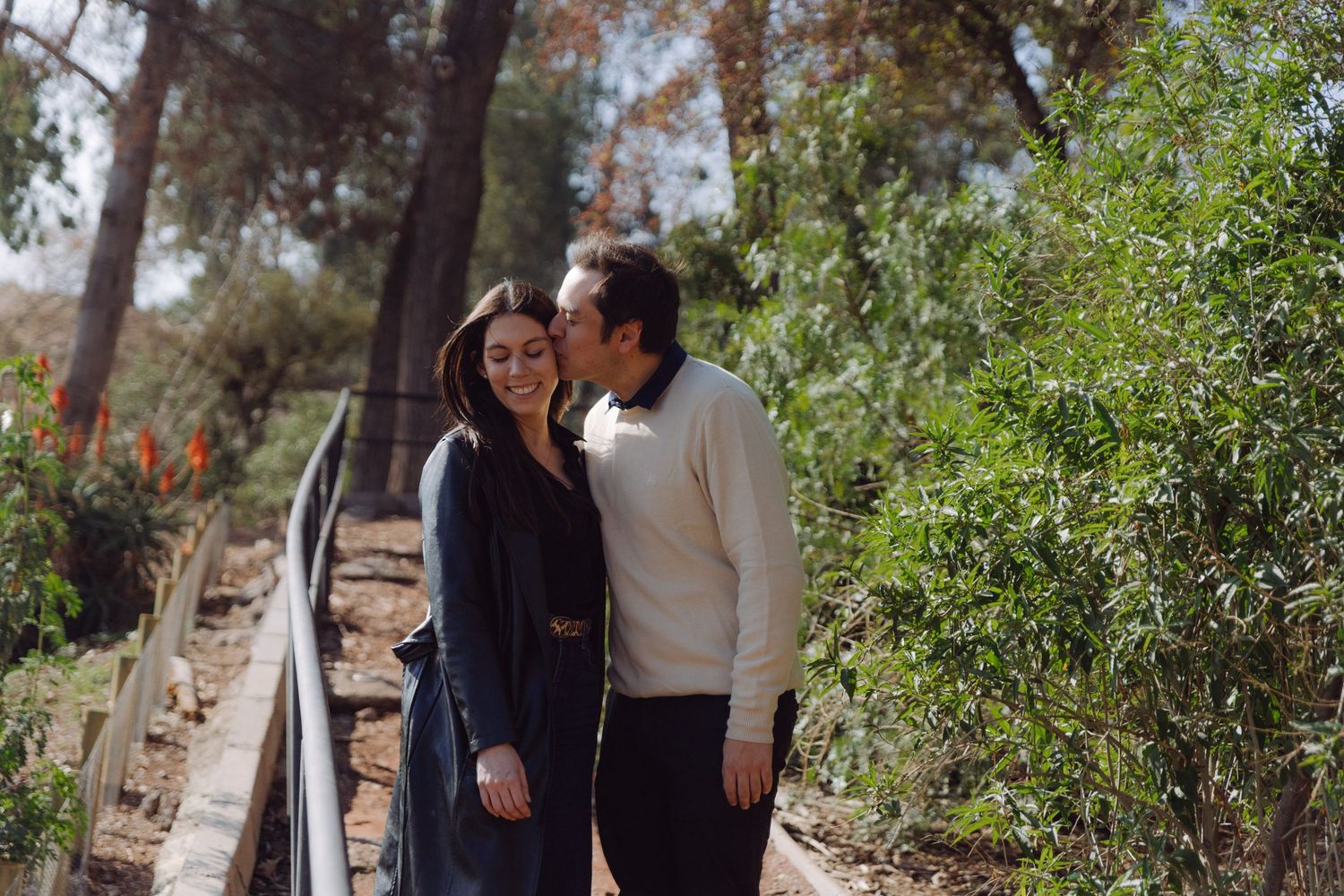 Man kissing woman on cheek as she smiles on a sunny garden path with green foliage.
