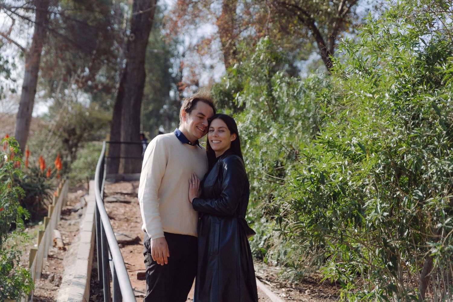 Smiling couple embracing on a garden path, woman in black leather coat and man in cream sweater.