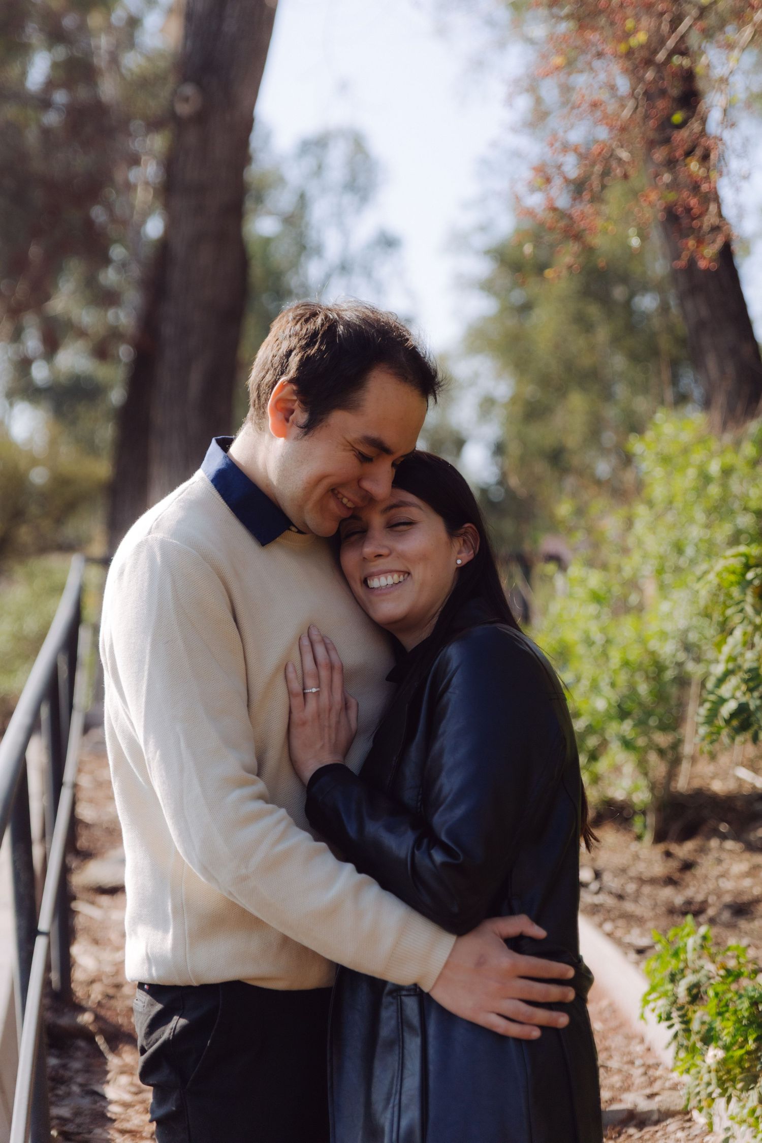 Woman smiling while hugging man on a sunny park path, showing engagement ring on her hand.