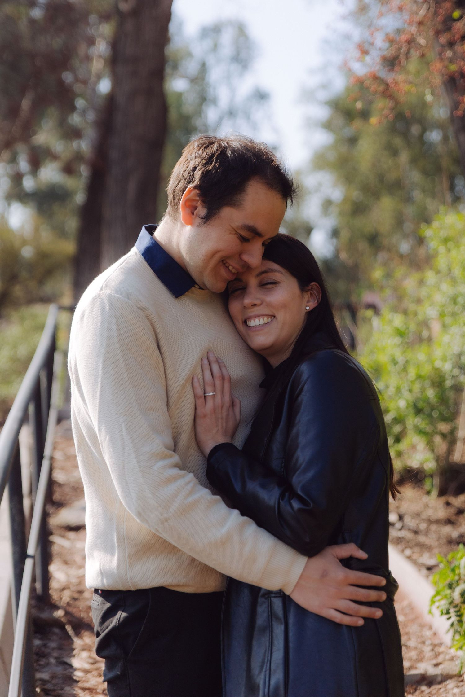 Happy couple embracing outdoors, woman displaying engagement ring while smiling in a sunlit park.