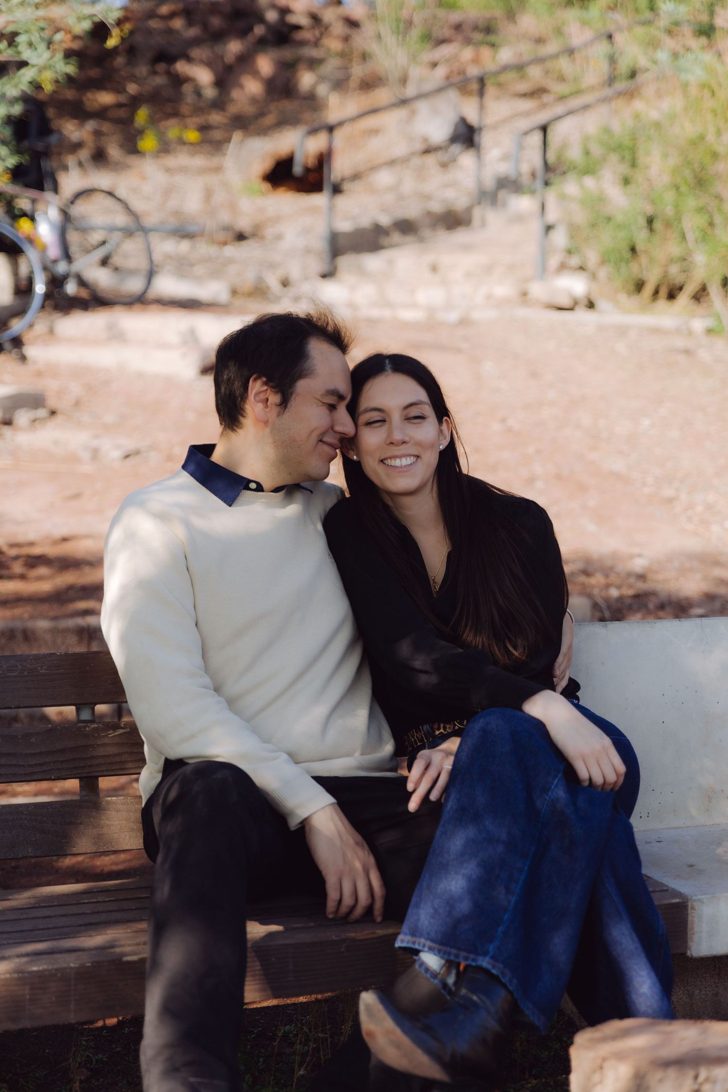 Smiling couple sitting together on a wooden bench in a sunny park, looking happy and relaxed.