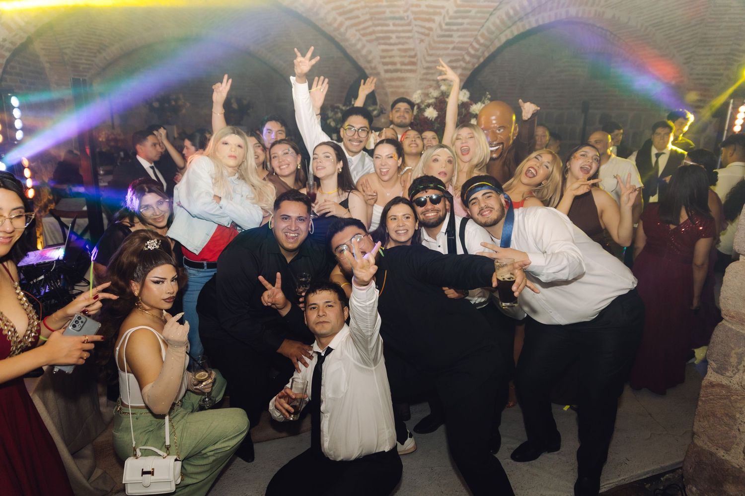 Large group of wedding guests dancing and celebrating together in a brick-vaulted venue with colorful party lights.