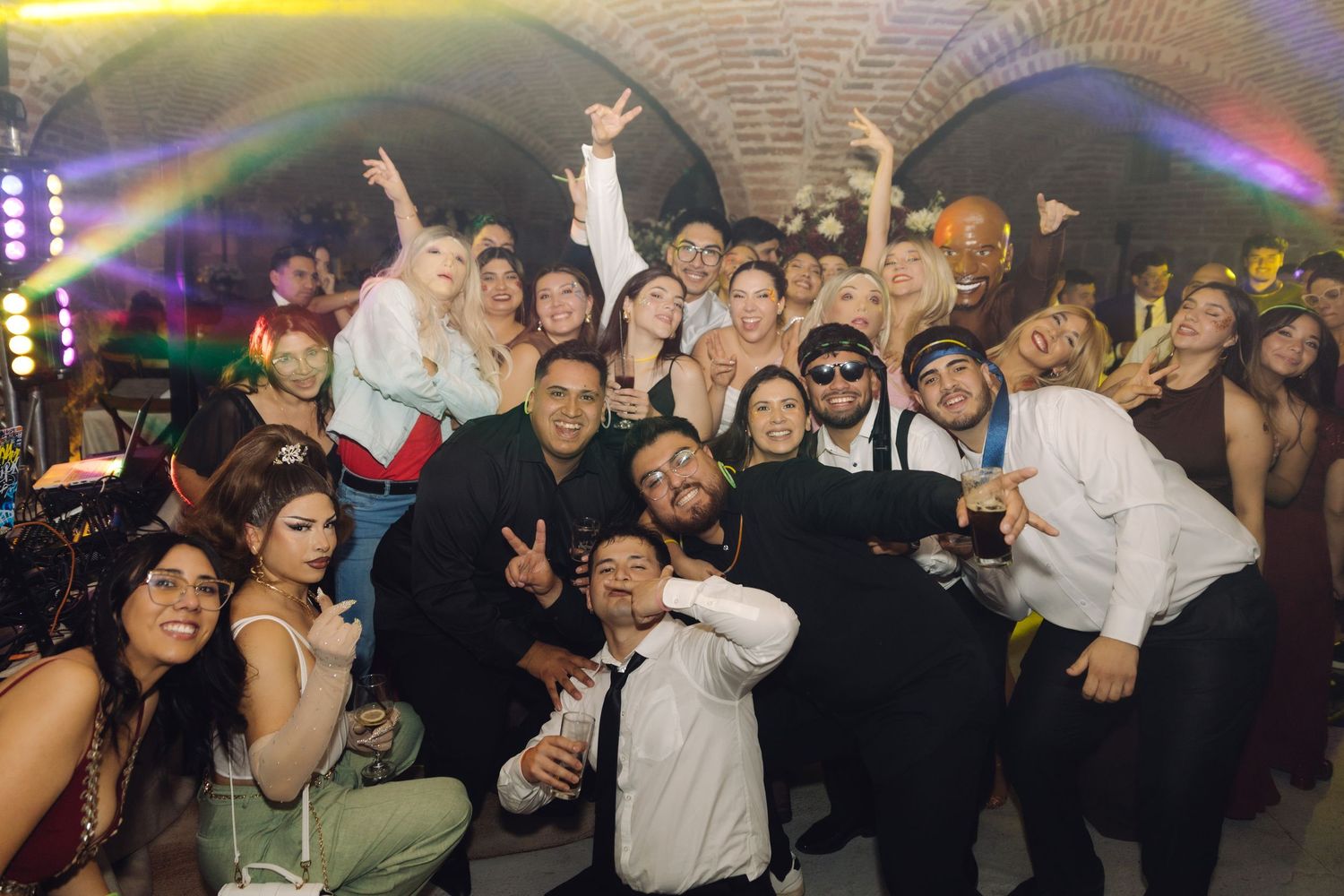 Joyful wedding party crowd posing for a group selfie under arched brick ceilings with vibrant lighting.