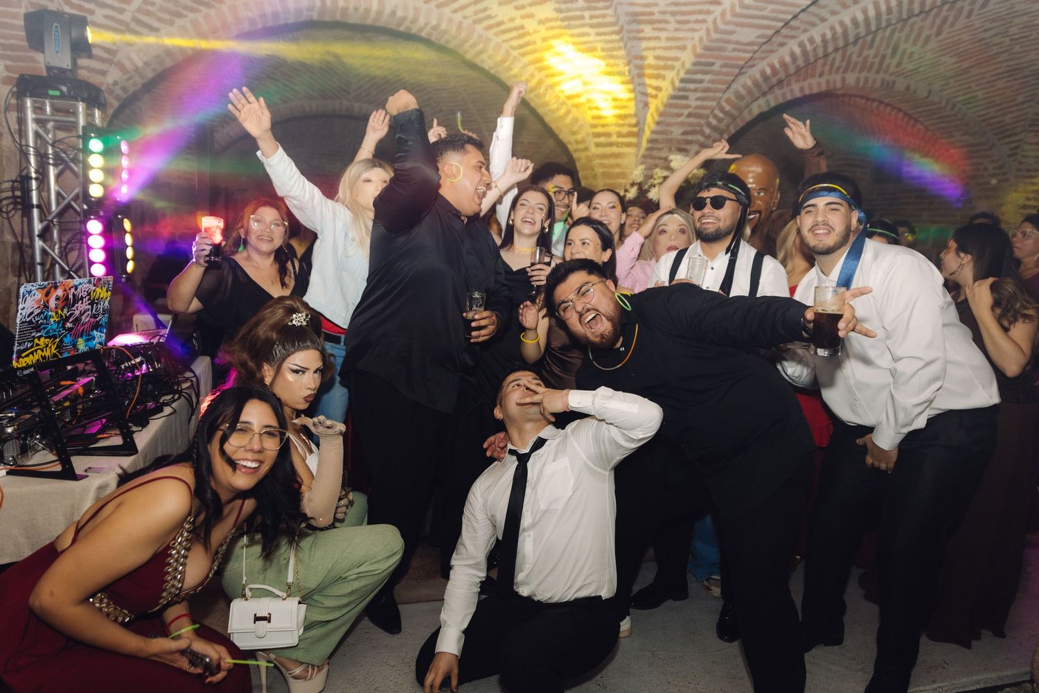 Excited wedding guests dancing and posing in a rustic brick-arched venue illuminated by colorful party lights.