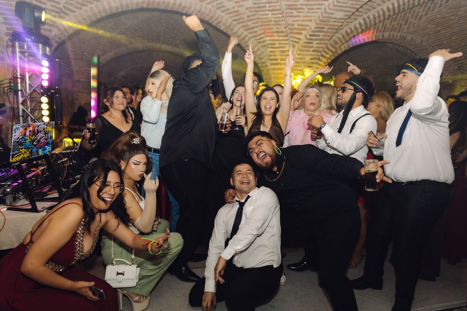 Wedding guests celebrating and dancing energetically in an elegant brick-vaulted reception venue with party lights.