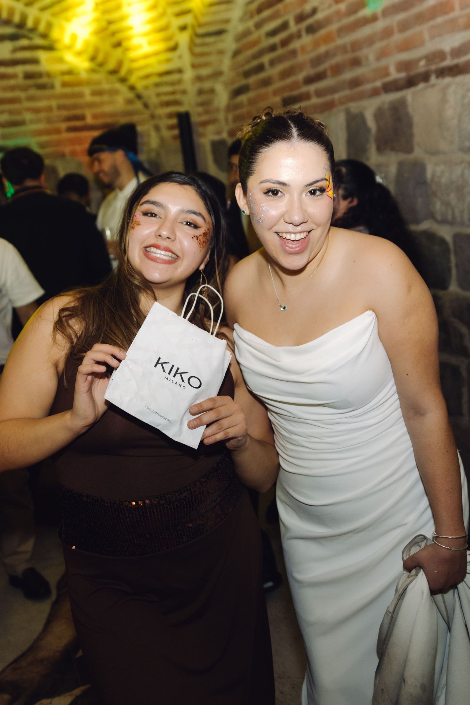 Two women smiling at a wedding reception, one in white bridal gown holding a KIKO branded bag.
