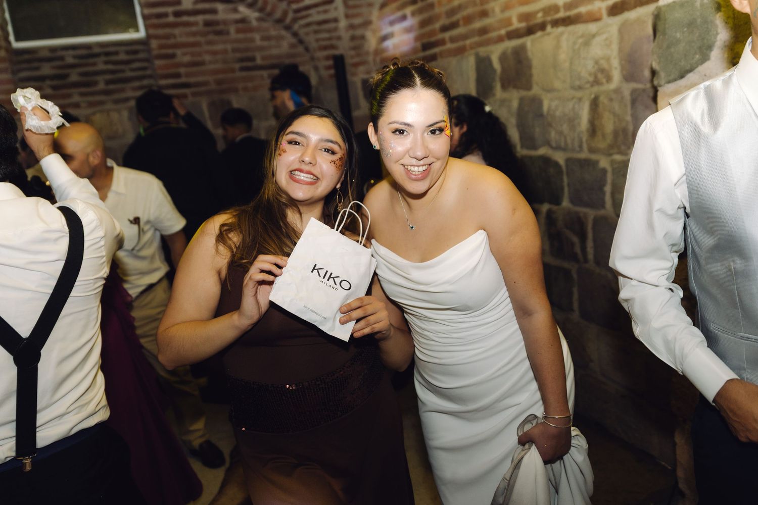 Bride in white strapless gown posing with a guest holding a KIKO shopping bag at a wedding reception.
