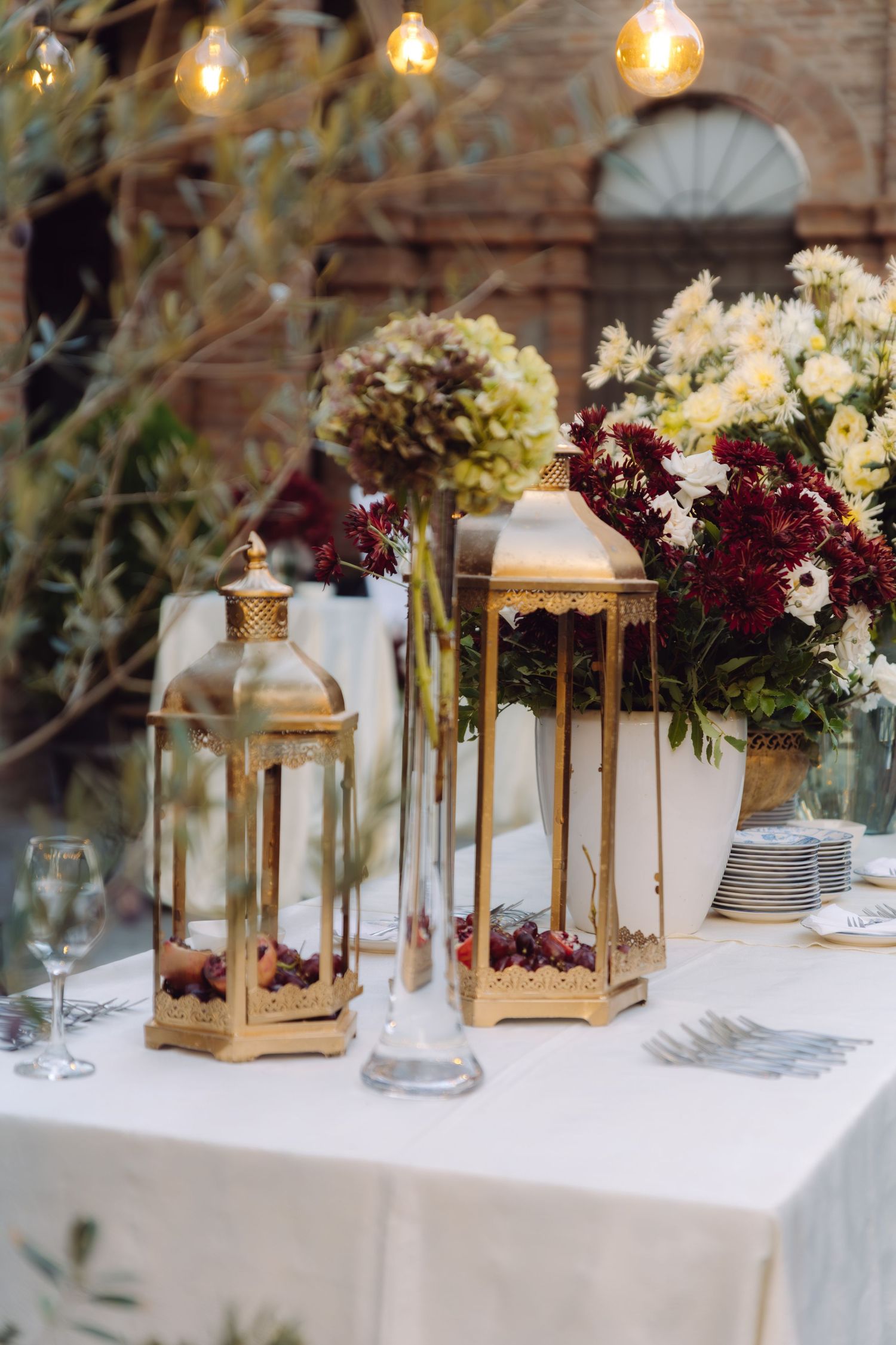 Gold decorative lanterns and lush white and burgundy floral centerpieces on a white-draped wedding reception table.