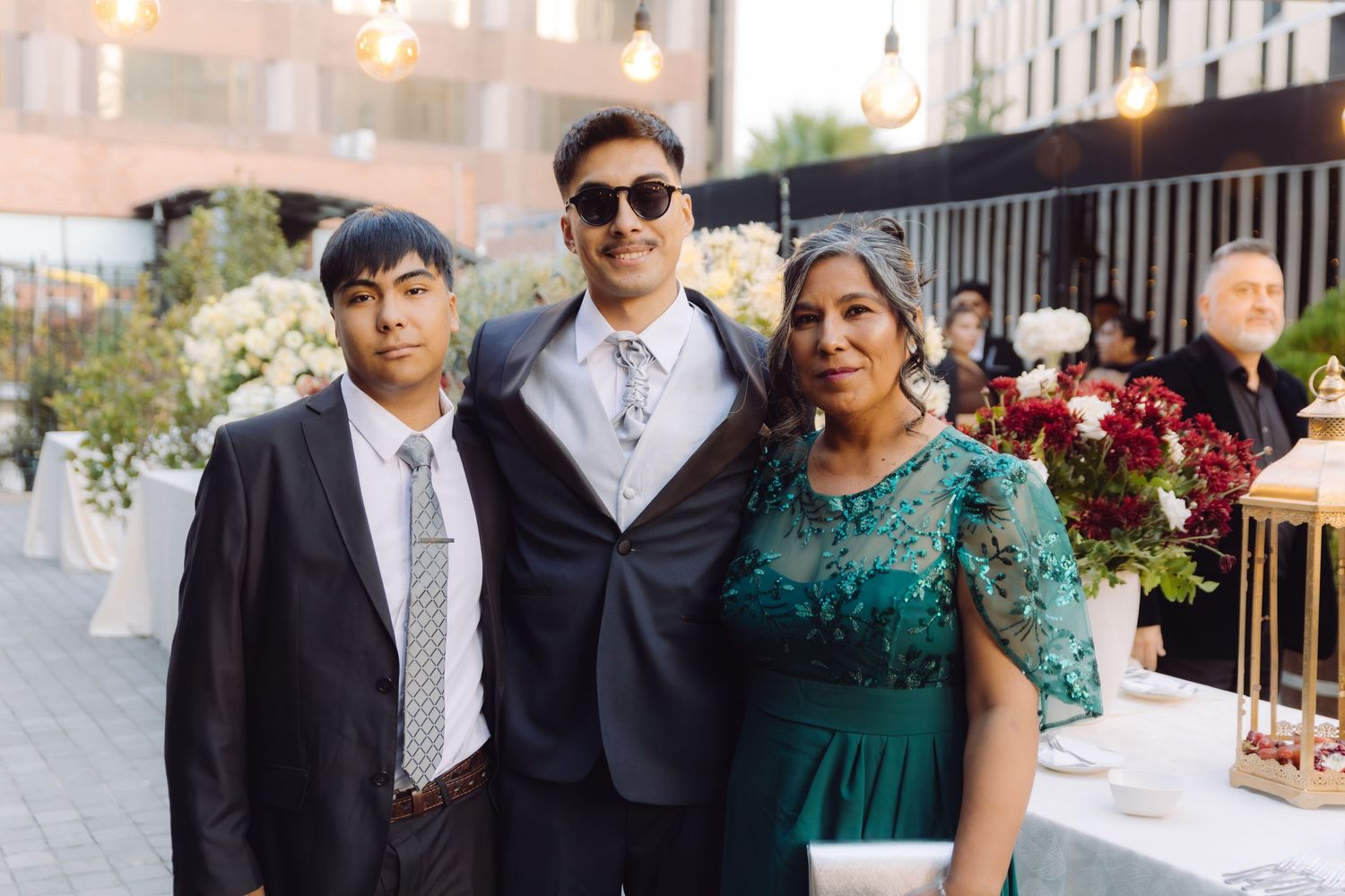Two young men and a woman in formal attire posing together at an outdoor wedding reception.