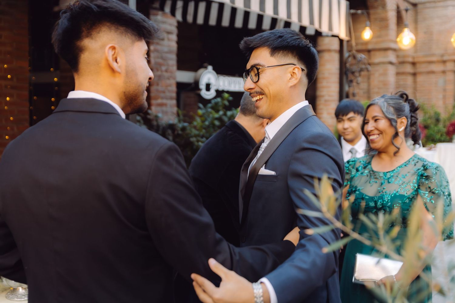 A groom in glasses and a cap greets guests at an outdoor wedding reception with warm lighting.