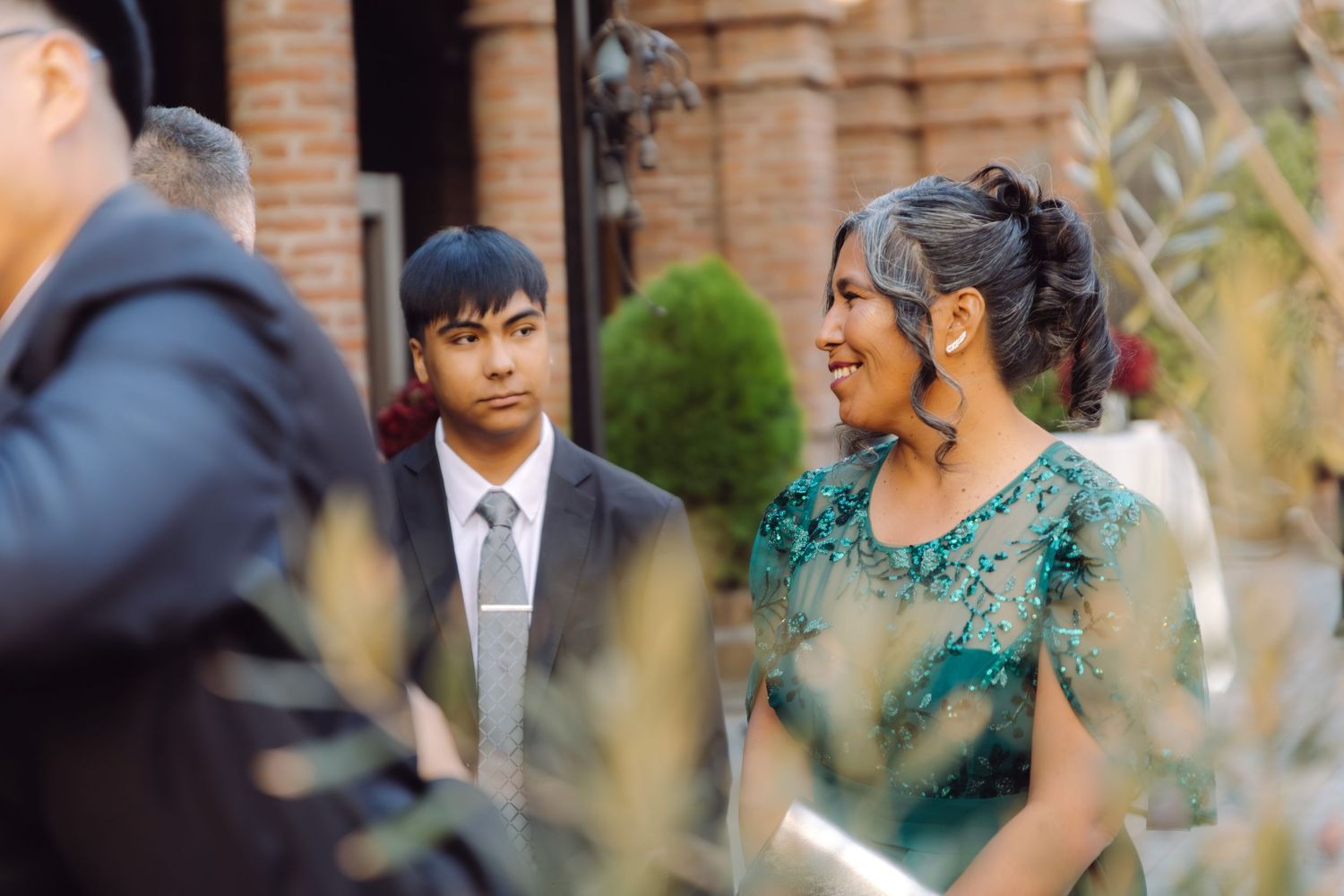 A young man in a suit and a woman in a green lace dress chat at an outdoor wedding venue.