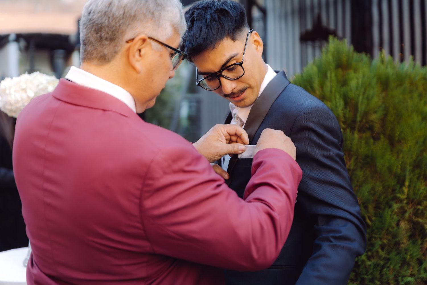 An older man in a red suit pins a boutonniere on a groom in a dark suit at a wedding.