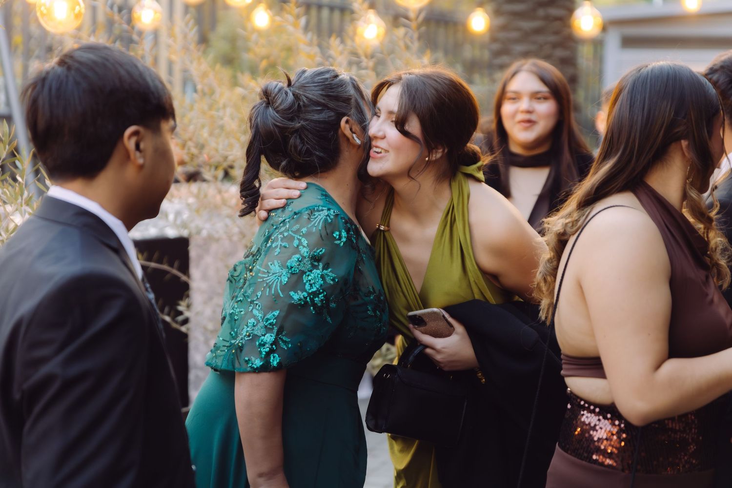 Wedding guests in colorful dresses embrace and greet each other at an outdoor evening reception.