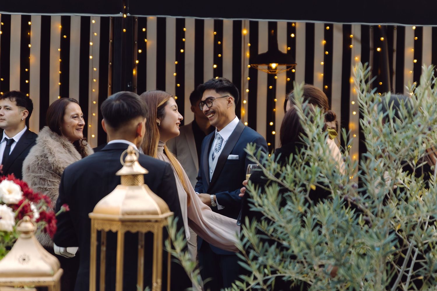 Guests in formal attire mingle at an outdoor wedding reception with string lights and greenery.