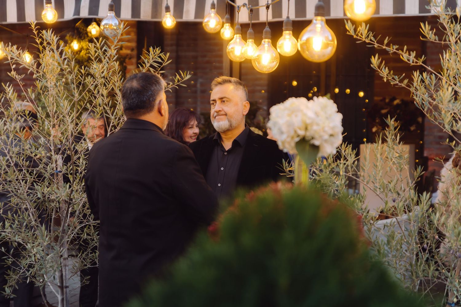 Two men in black suits converse at an outdoor wedding venue decorated with string lights and plants.