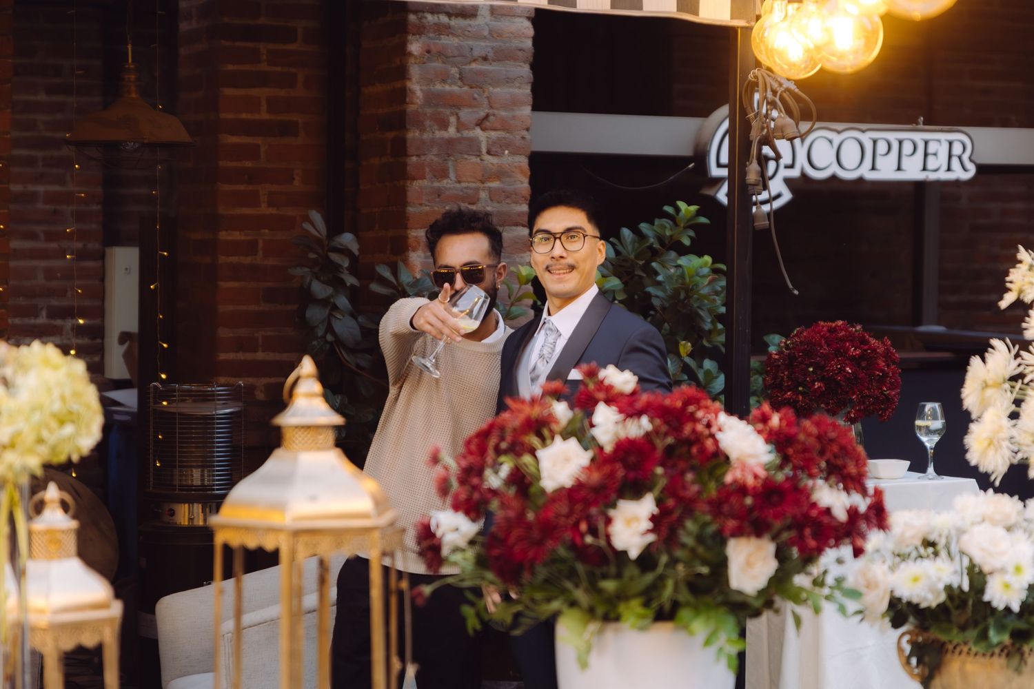 Two men in suits smile and take a photo near red and white floral arrangements at a wedding.