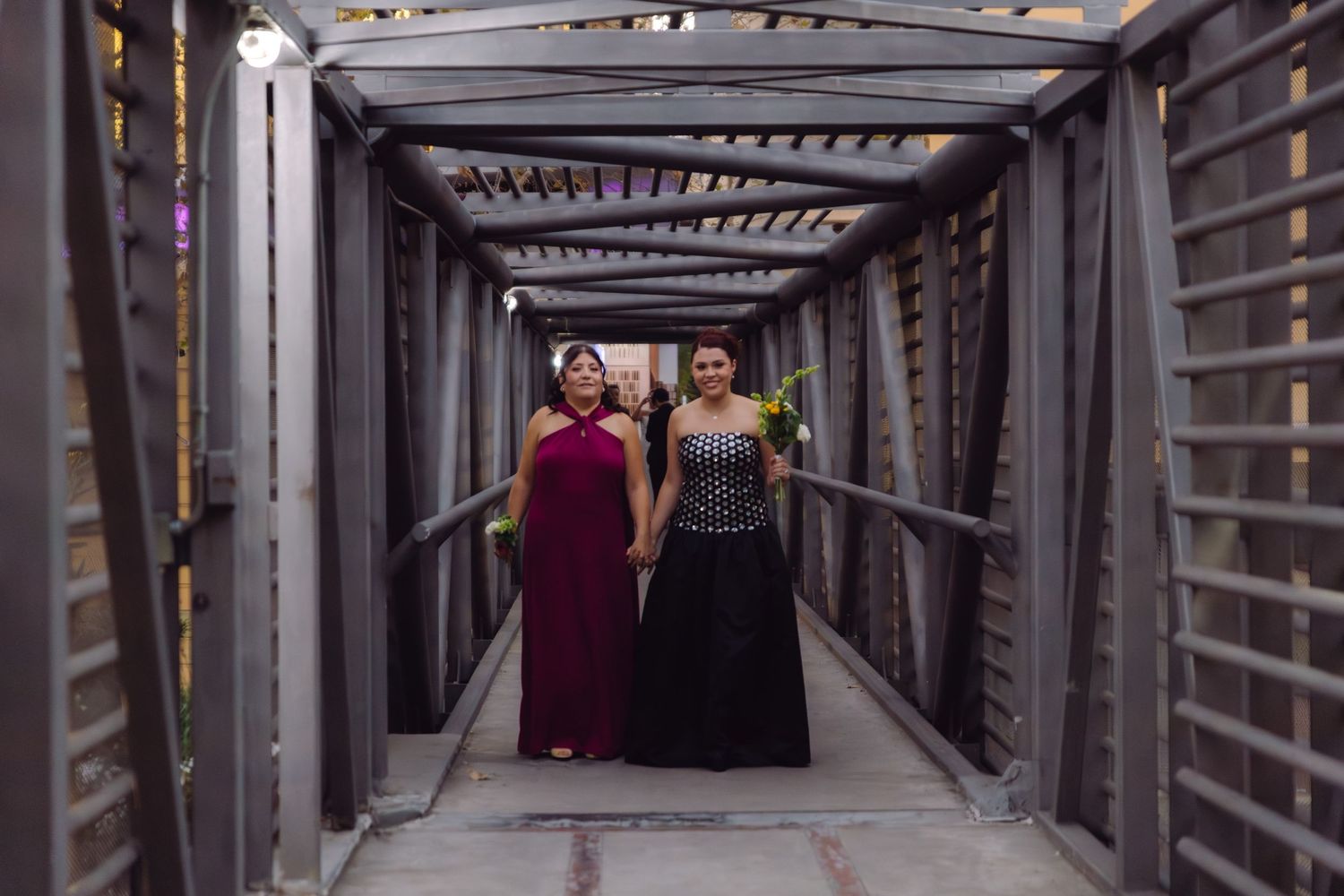 Two women in formal gowns pose together on a decorated walkway at a wedding event.
