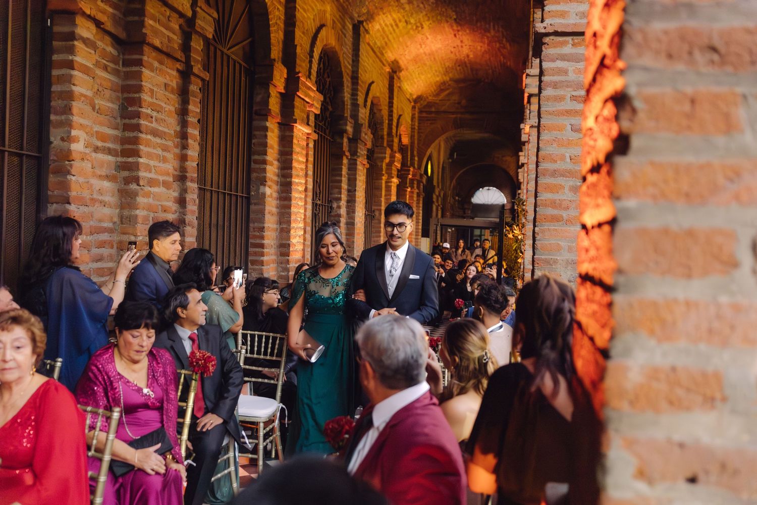 Wedding guests walk down an arched brick corridor aisle during an outdoor ceremony.