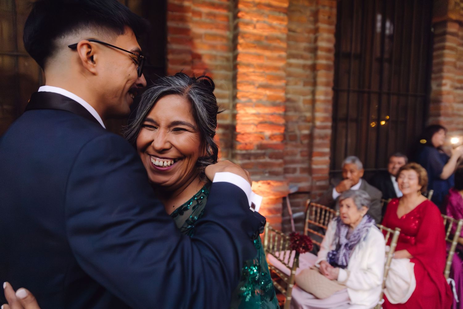 Bride smiles joyfully while embracing a guest in a green dress at a wedding ceremony.