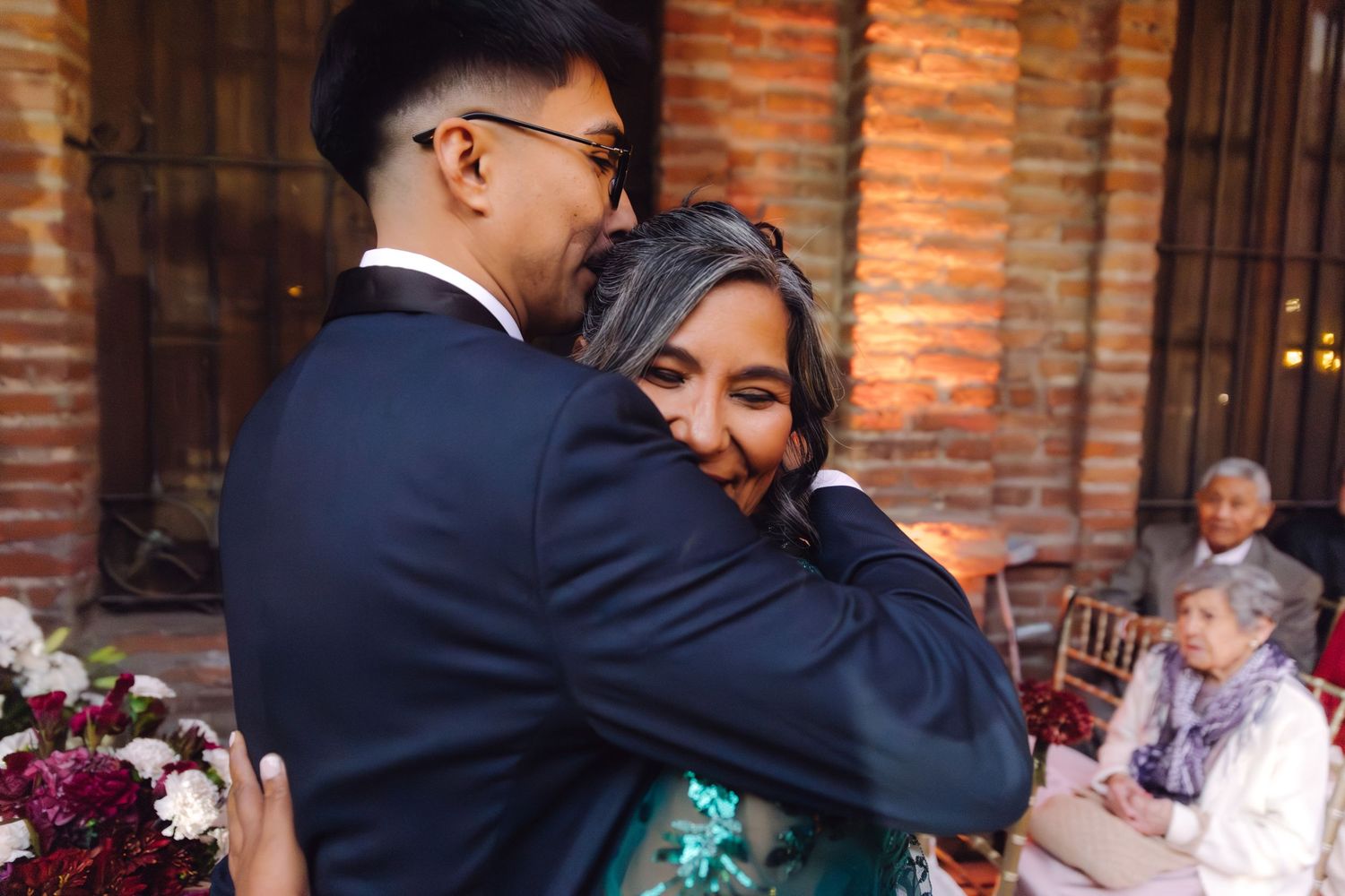 Emotional wedding moment as bride hugs a guest in a teal dress near brick archways.