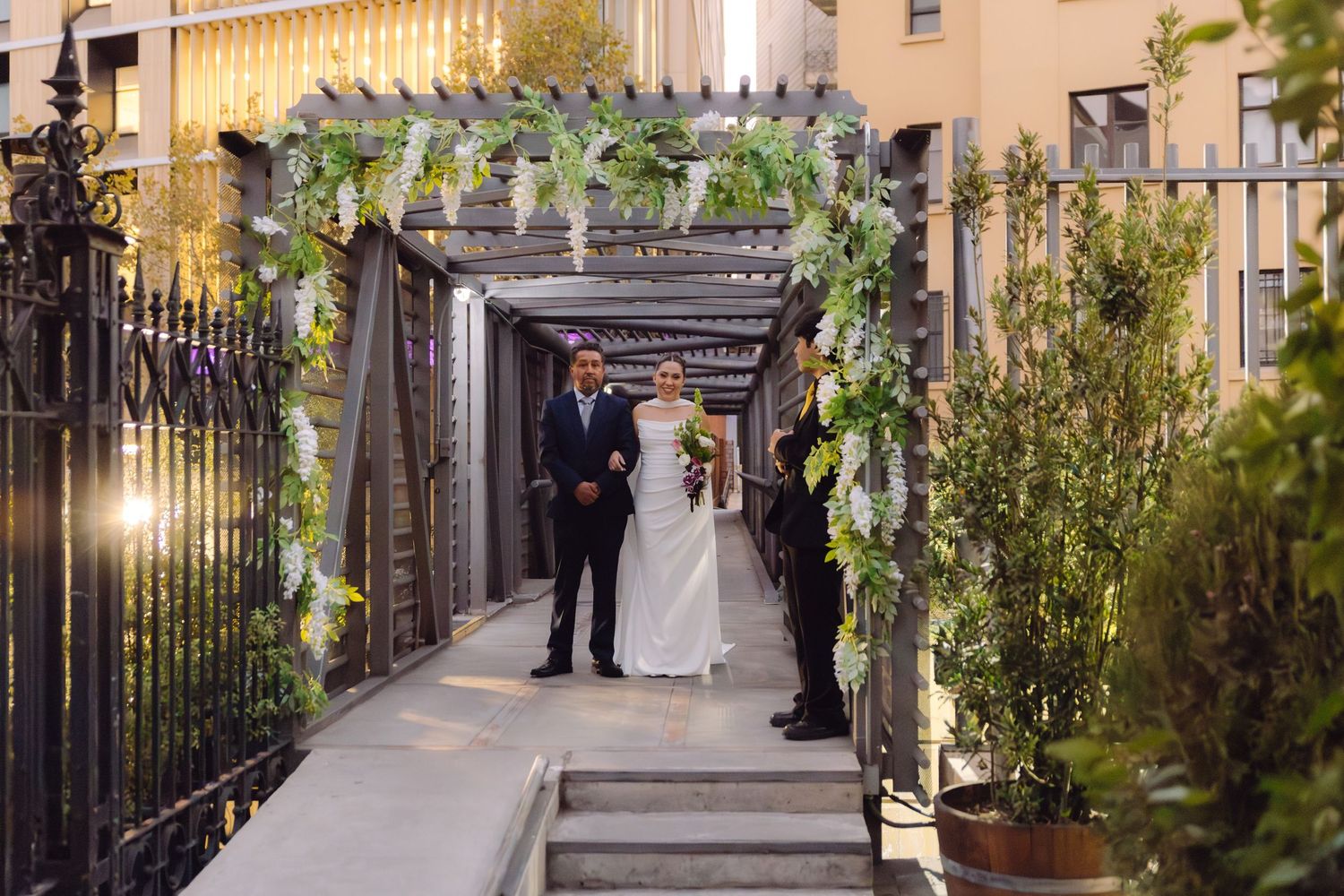Bride and escort walk under a floral pergola arch at an elegant outdoor wedding venue.