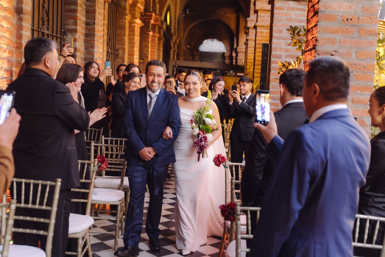 Bride and escort walk down the aisle at a wedding ceremony in a brick archway venue.