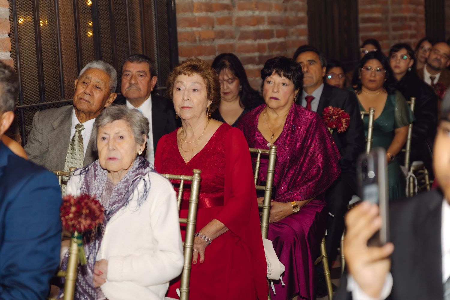 Wedding guests in formal attire watch the ceremony from gold chiavari chairs indoors.