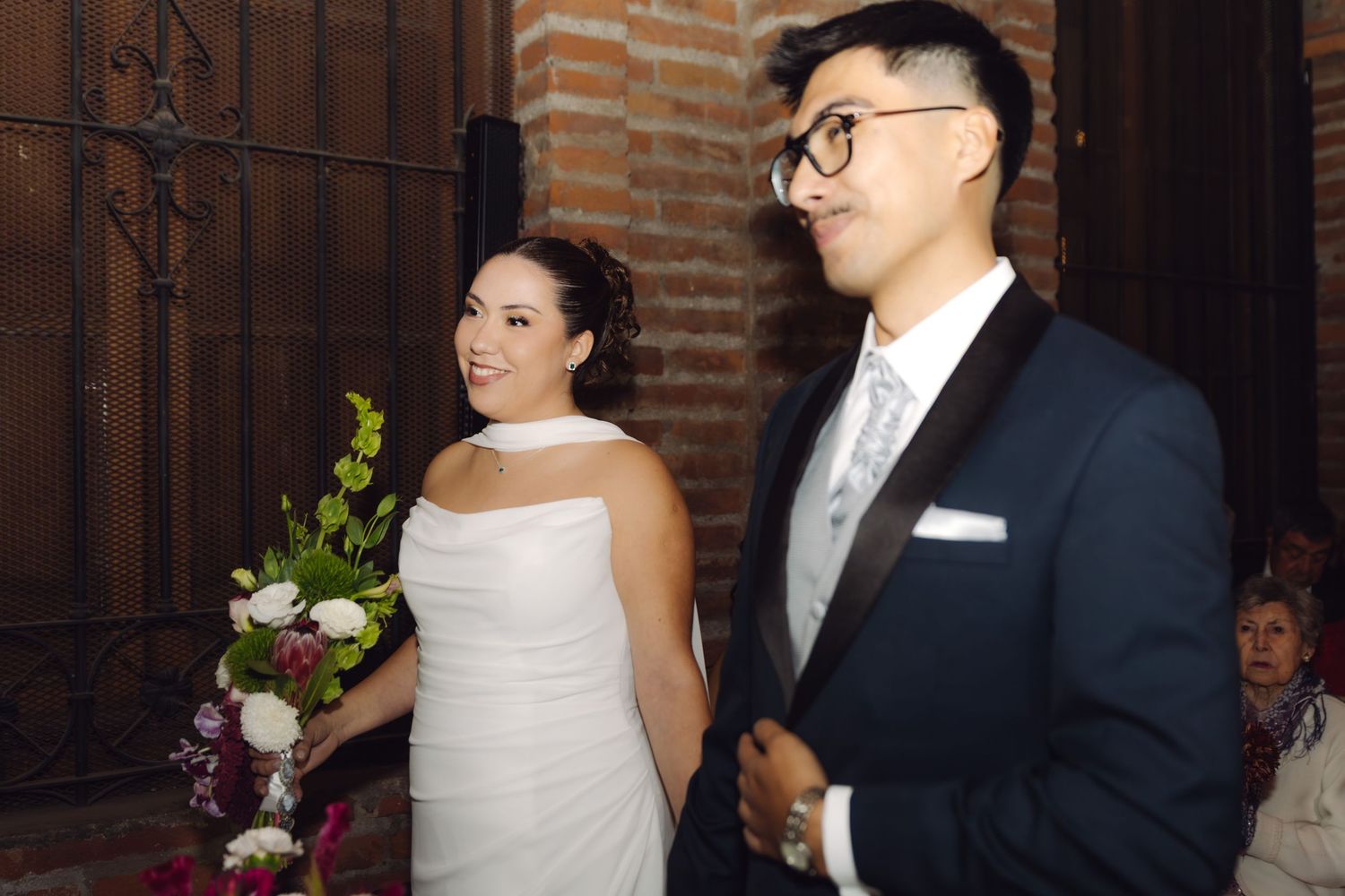 Smiling bride in white gown and groom in navy suit stand together at indoor wedding ceremony.