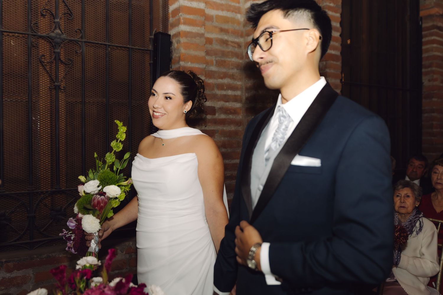 Bride in white dress holding bouquet stands with groom in navy tuxedo at wedding ceremony.