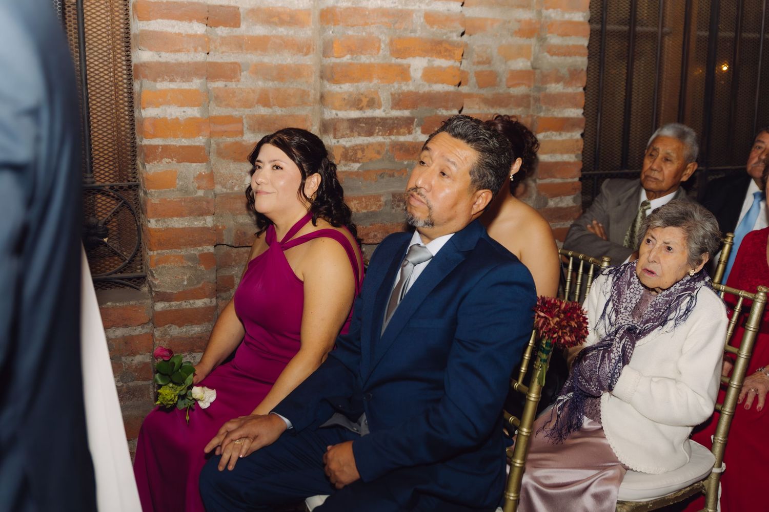 Wedding guests in formal attire seated on chairs watching ceremony in brick-walled venue.