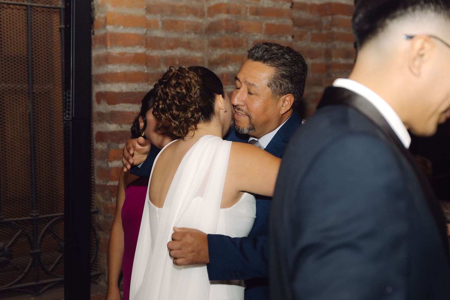 Bride in white dress shares warm embrace with older man in navy suit at wedding reception.