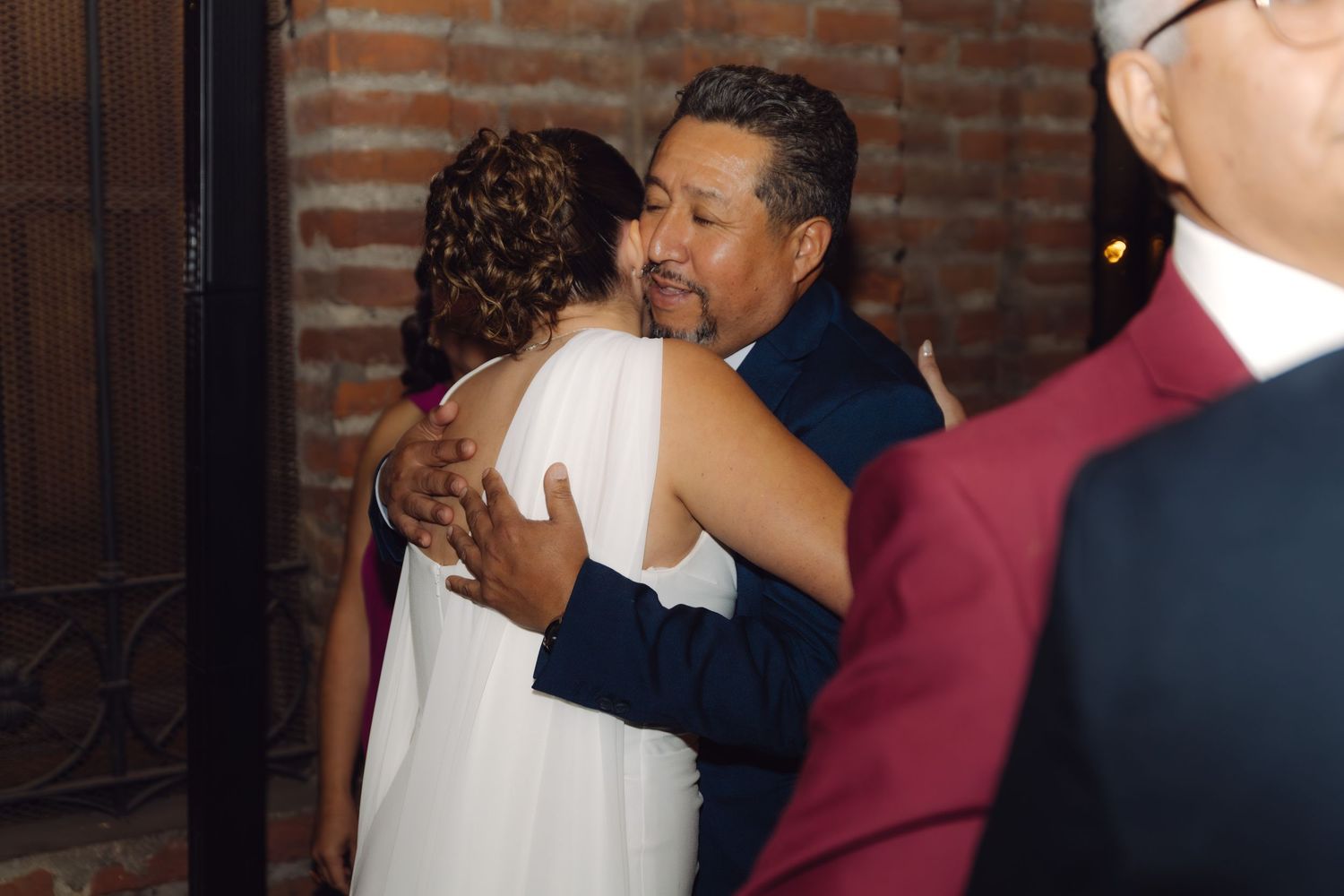 Bride in white dress embraces older man in navy suit warmly at wedding reception.