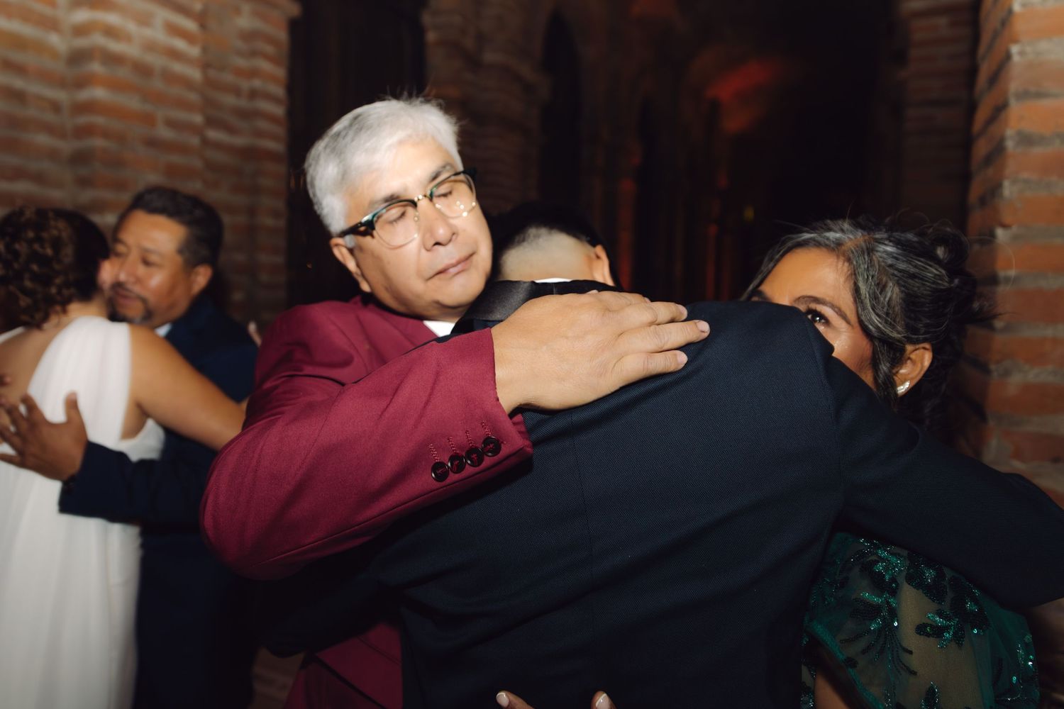 Wedding guests share warm embraces during joyful celebration in rustic brick-walled venue.