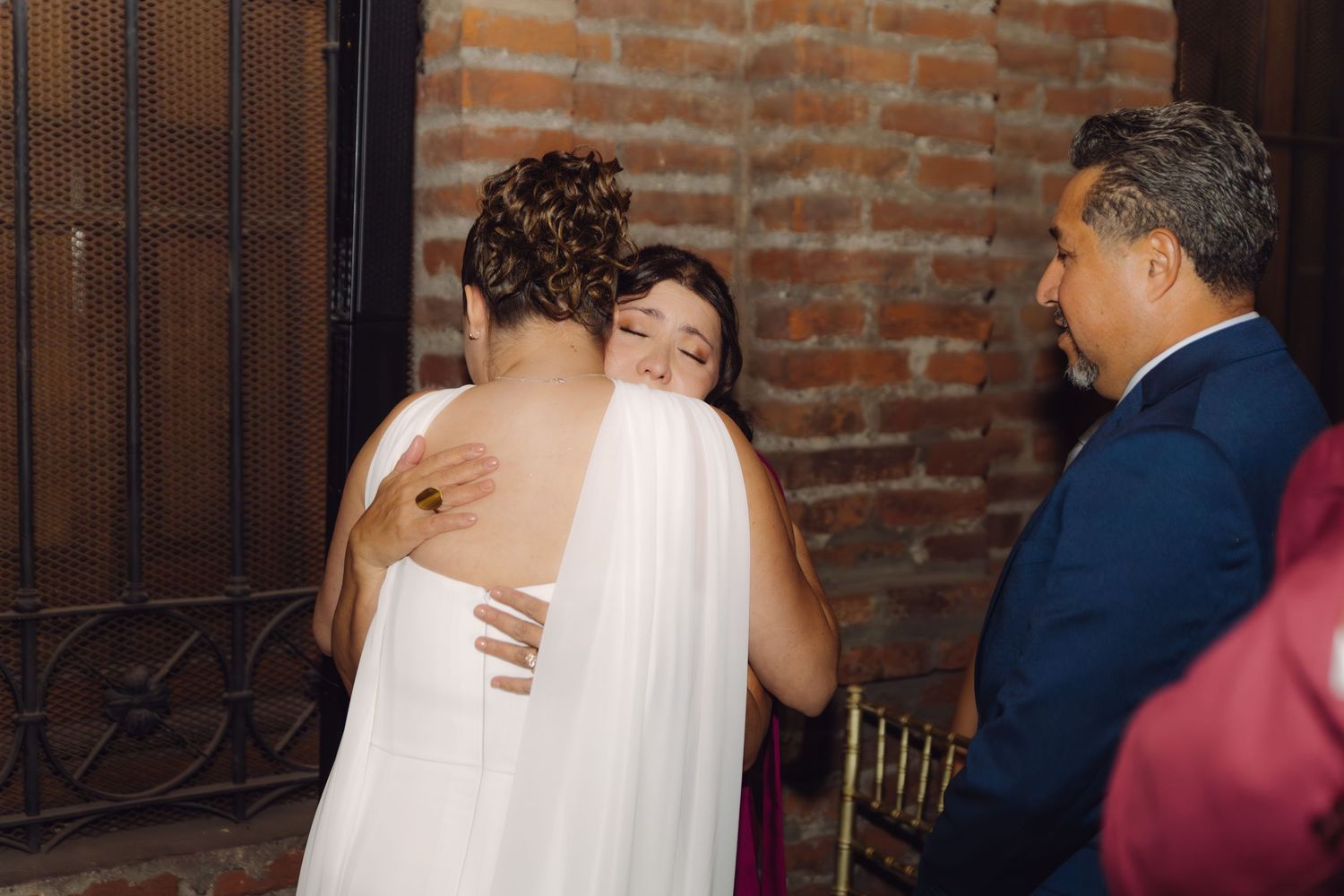 Bride in white gown shares emotional hug with smiling female guest at wedding reception.