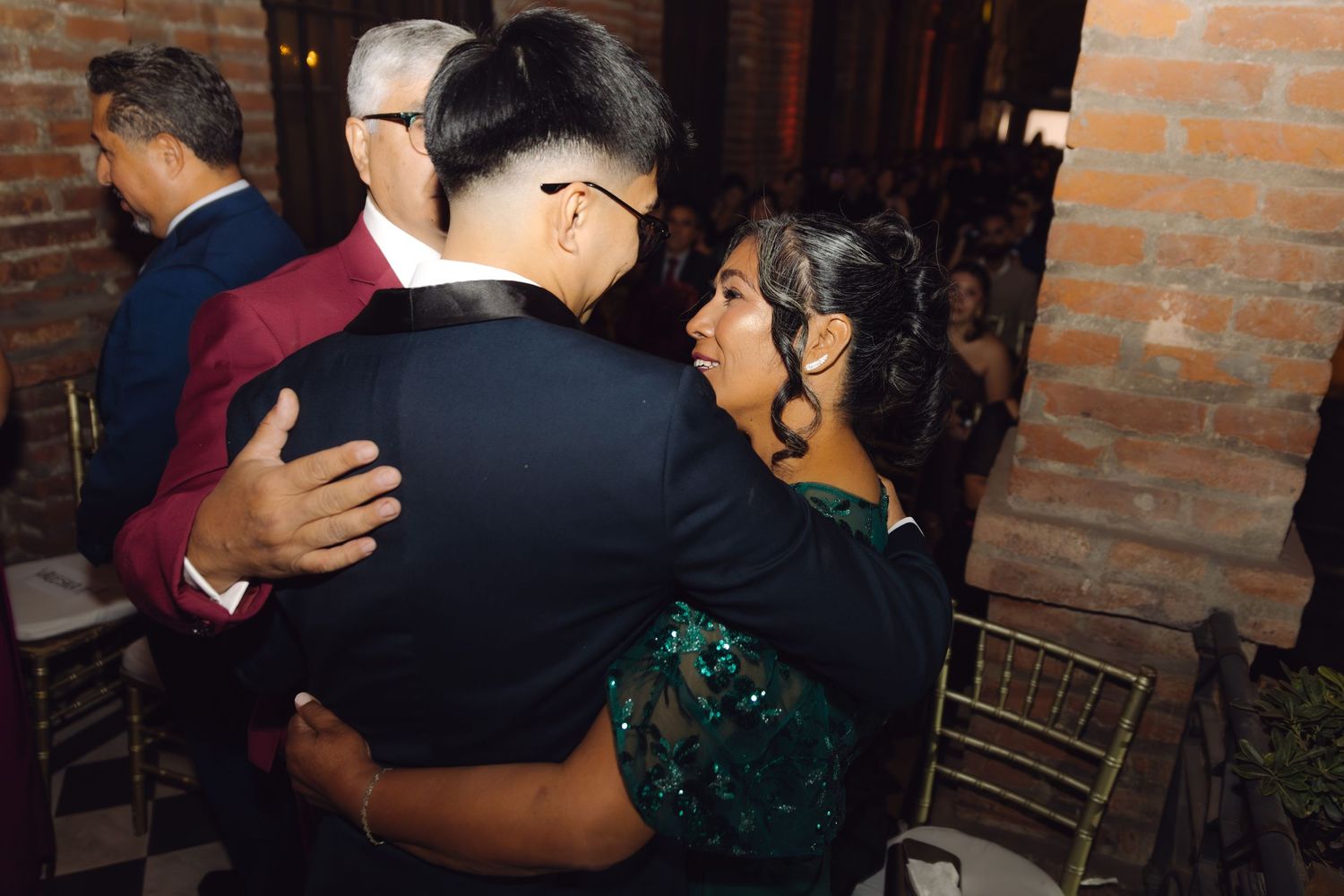 Groom in black suit shares warm embrace with woman in green sequin dress at reception.