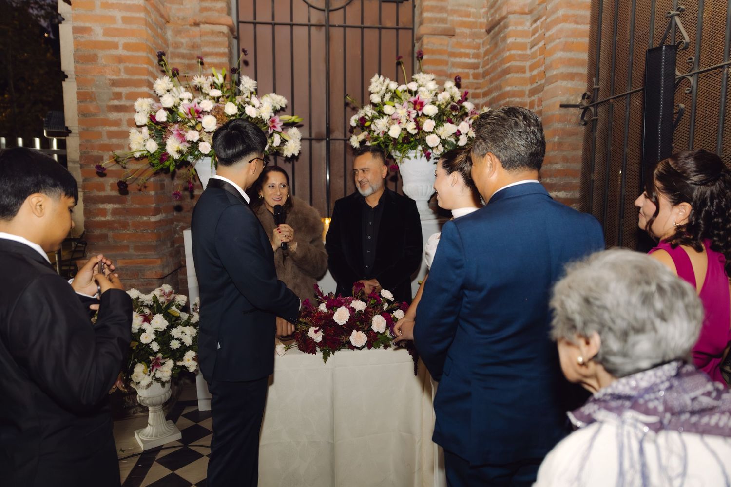 Wedding ceremony at floral-decorated altar with couple and officiant in rustic brick venue.