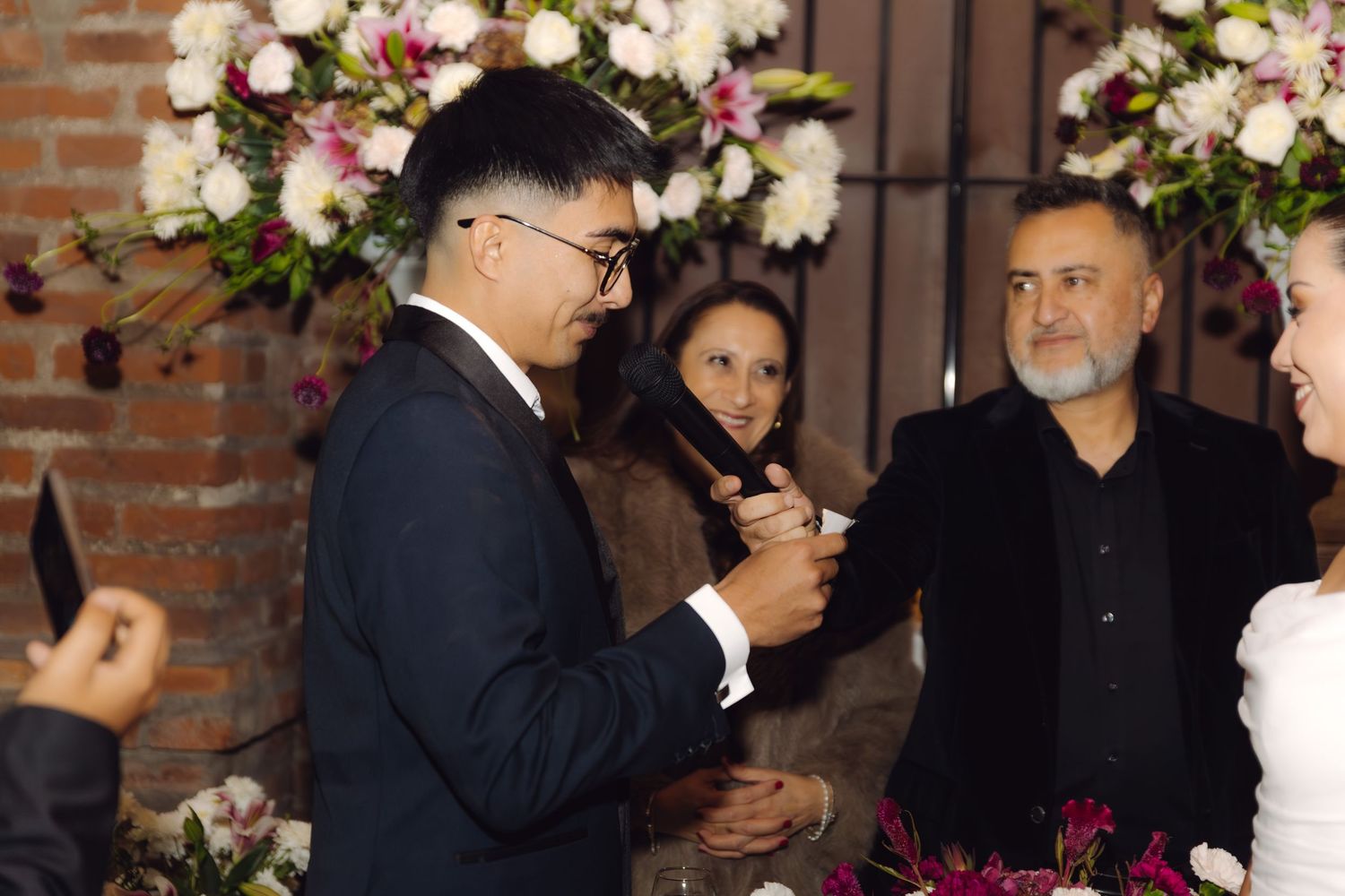 Groom in navy suit reading vows during wedding ceremony with floral decorations and guests nearby.