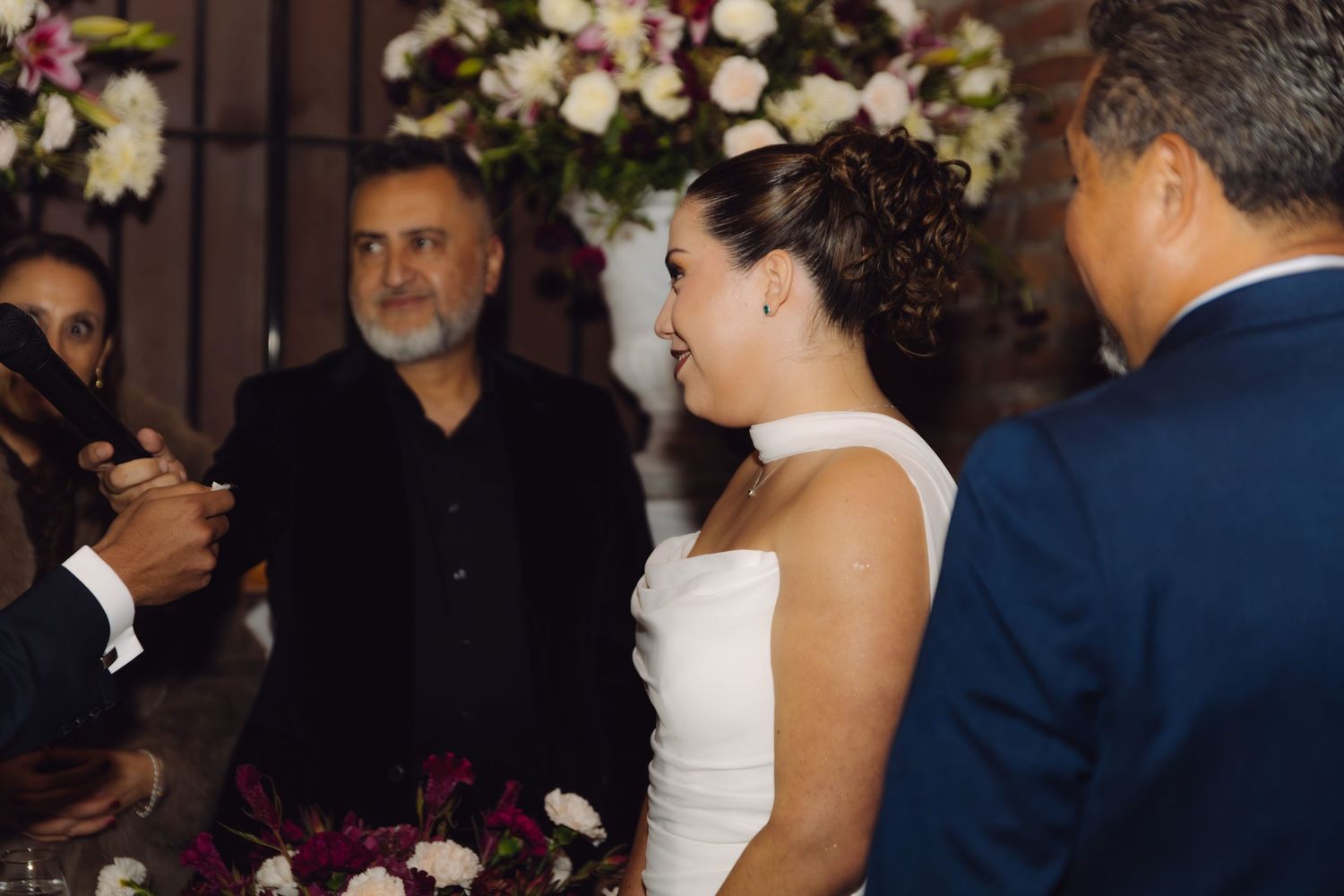 Bride in white gown smiling during wedding ceremony with officiant and floral arrangements in background.