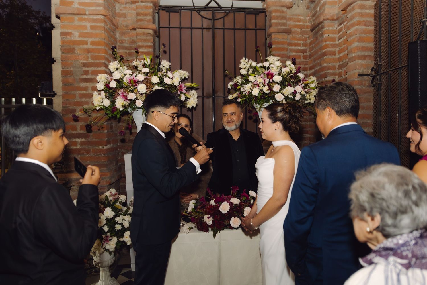 Couple exchanging vows at outdoor wedding ceremony with brick wall backdrop and elegant floral arrangements.